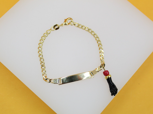 A gold-plated ID bracelet featuring a curb chain and a figa charm with a red bead rests on a white display surface.