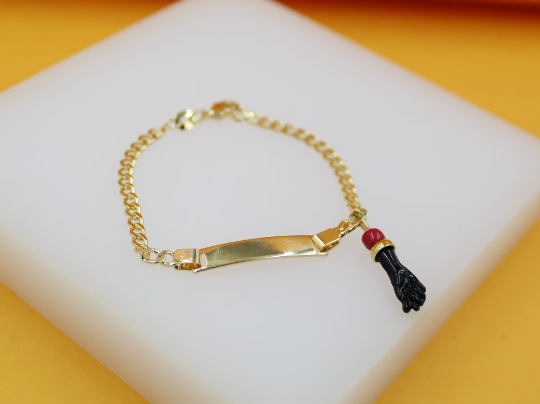 A gold-plated identity bracelet rests on a white display stand, featuring a blank ID plate and a figa charm with a red bead hanging from the chain.