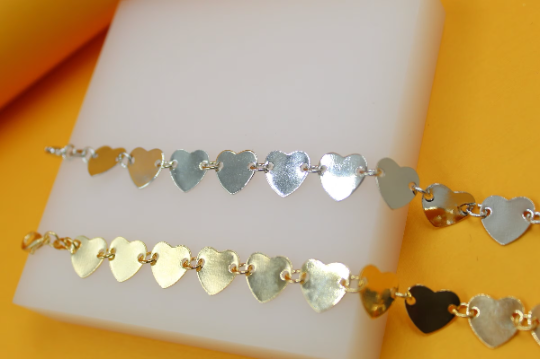 Two delicate gold heart-shaped charm bracelets rest elegantly on a white display block against a vibrant yellow background.
