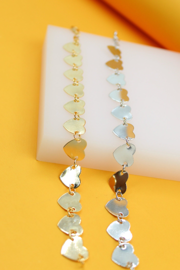 Two delicate heart-link chains, one in gold and one in silver, rest on a white display block against a vibrant yellow background.