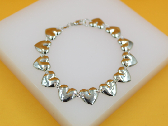 A silver heart-linked charm bracelet rests on a white display block against a yellow background.