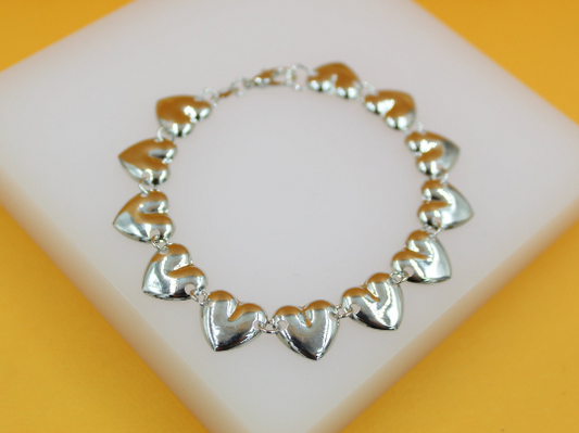 A silver heart-linked charm bracelet rests on a white display block against a yellow background.