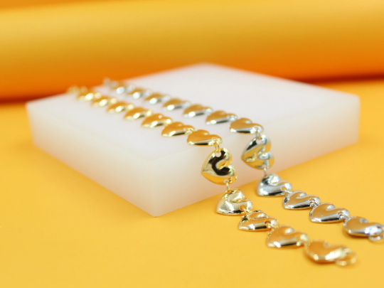 Two shiny heart-link bracelets, one in gold and one in silver, rest elegantly on a white display block against a vibrant yellow background.