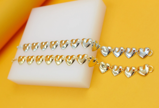 Two delicate metallic heart-link bracelets, one silver and one gold, rest neatly on a white display block against a vibrant yellow background.