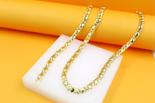 A shimmering gold-plated hug and kiss tennis necklace rests gracefully on a white display block against a vibrant yellow background.