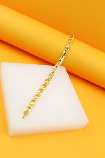 A delicate gold-plated X and O tennis bracelet rests diagonally on a white pedestal against a vibrant orange background.