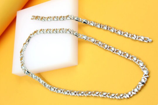 A matching silver heart and cross tennis necklace and bracelet set rests on a white display block against a vibrant yellow background.