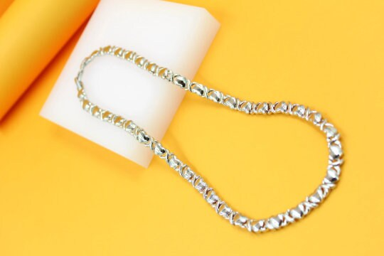 A polished silver XO link necklace rests gracefully on a white display block against a vibrant yellow background.