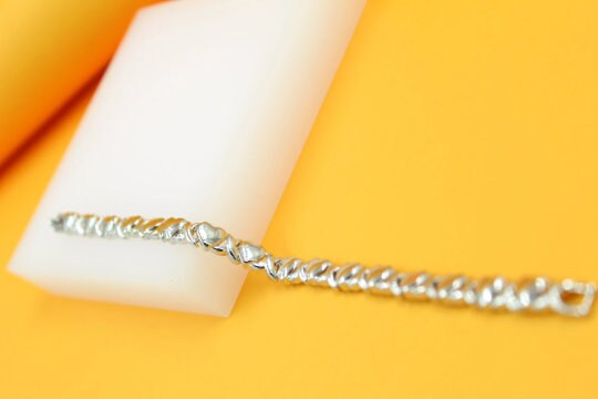 A silver heart-link tennis bracelet rests diagonally across a white display block against a vibrant yellow background.