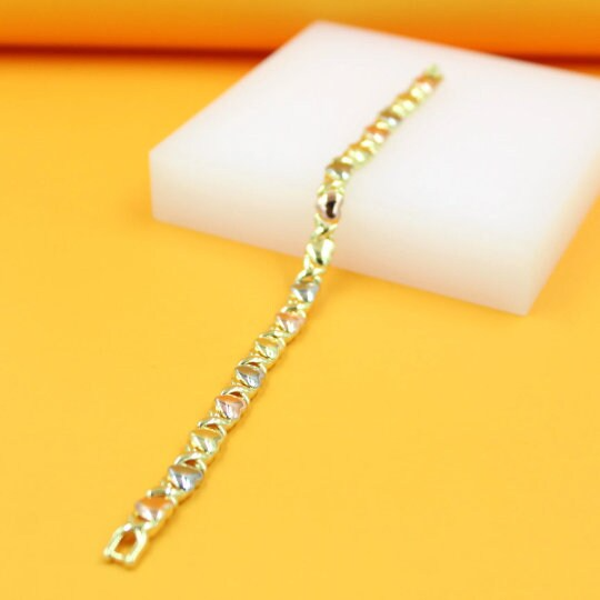 A delicate tri-color gold heart link bracelet rests diagonally on a white display block against a vibrant yellow background.