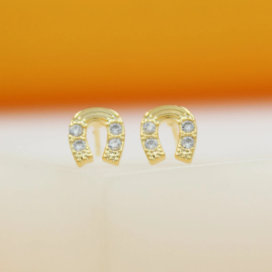 A pair of gold horseshoe stud earrings sparkle with embedded cubic zirconia stones against a warm orange backdrop.