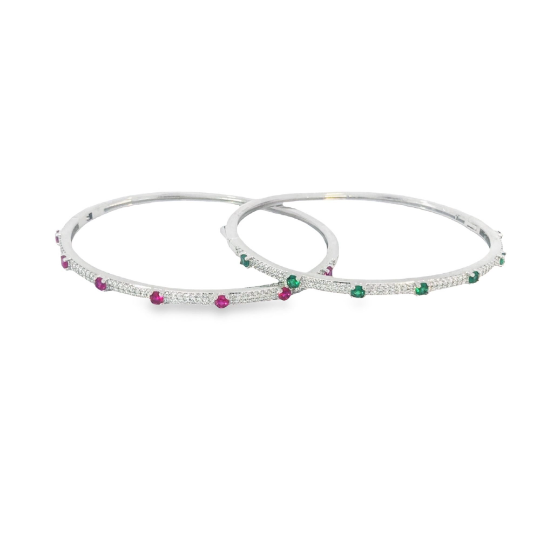 Two silver diamond bangles encrusted with sparkling pave diamonds and accented with vibrant red ruby and green emerald gemstones rest side by side on a clean white surface.