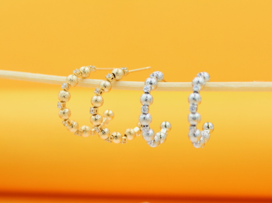 Two pairs of gold and silver textured bead hoop earrings hang delicately from a wooden stick against a warm yellow background.