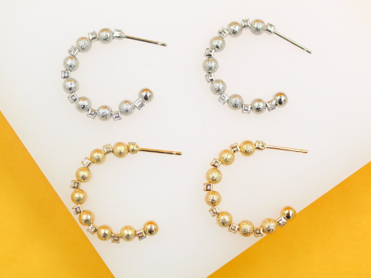Two pairs of textured gold and silver hoop earrings featuring alternating beaded spheres and crystal accents rest on a white display surface against a vibrant yellow background.