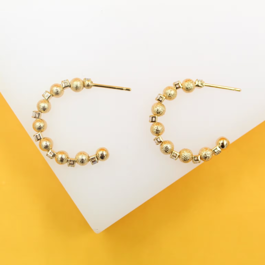 A pair of textured gold bead hoop earrings rests against a white and yellow background.