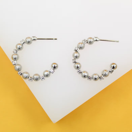 A pair of sterling silver hoop earrings features textured spheres and small square spacer elements against a white and yellow background.