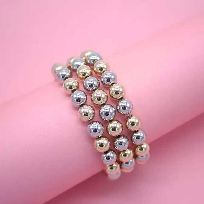Three beaded bracelets featuring alternating gold and silver metallic spheres rest gracefully on a soft pink surface.