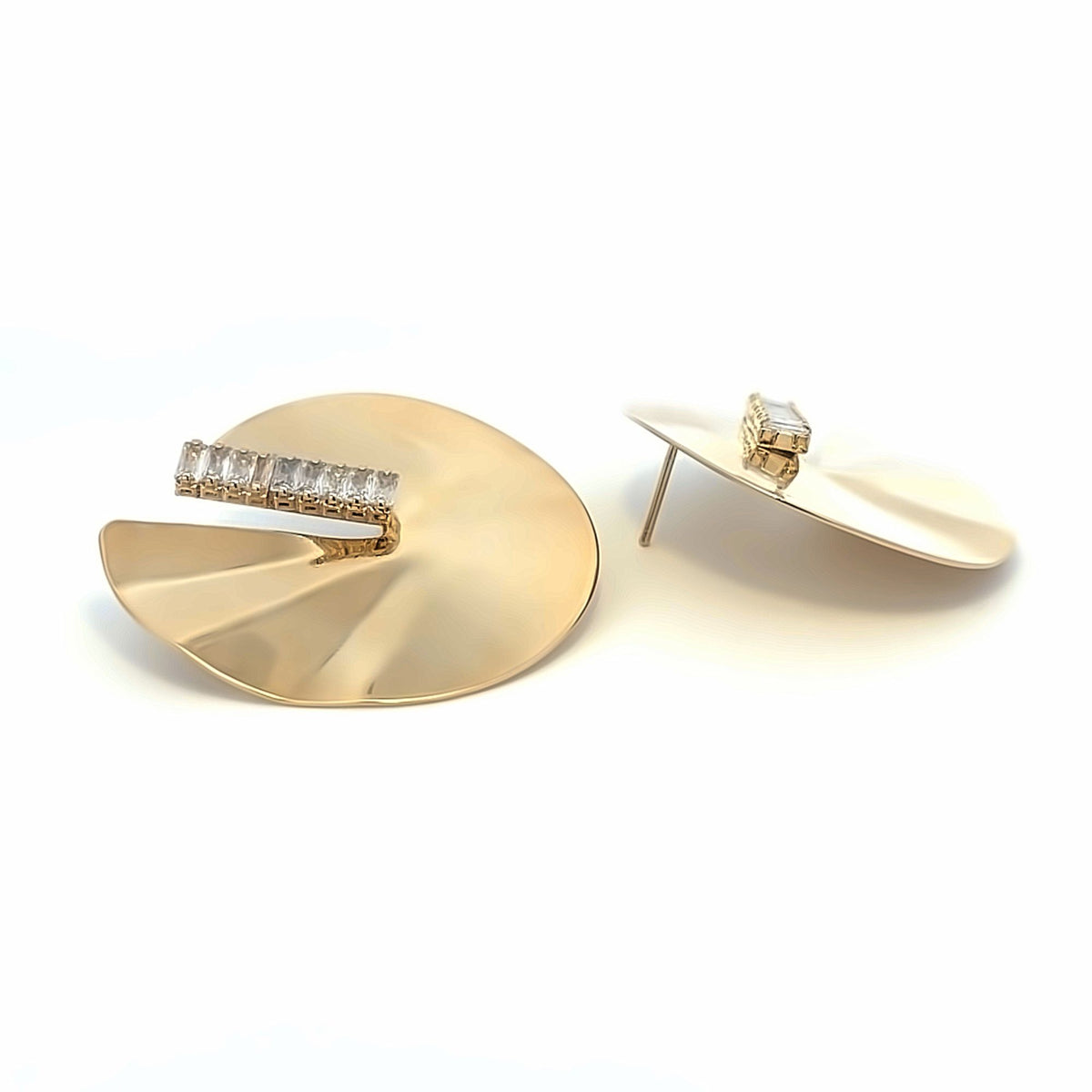 A pair of modern gold disc earrings featuring a delicate row of sparkling baguette-cut crystals rests on a clean white surface.