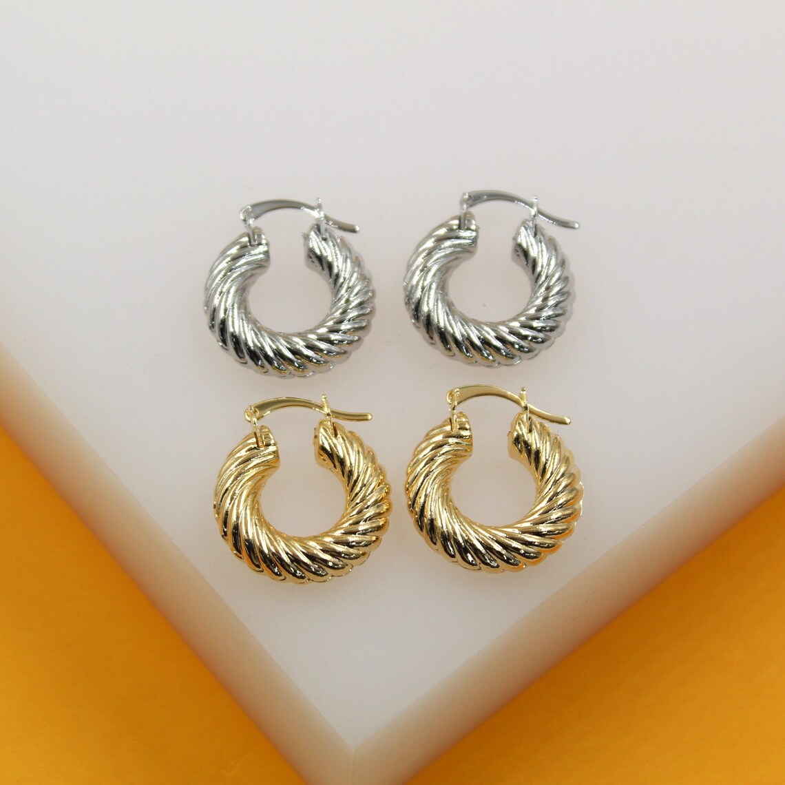A pair of silver and a pair of gold twisted dome hoop earrings rest side by side on a white display stand.