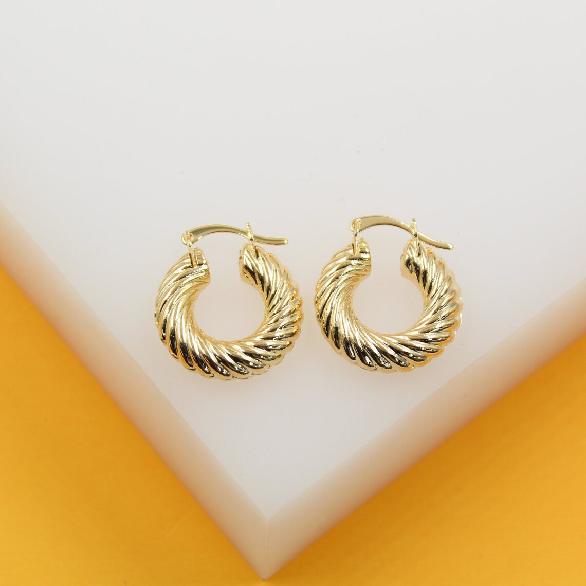 A pair of shiny gold twisted hoop earrings rests on a white and yellow display stand.