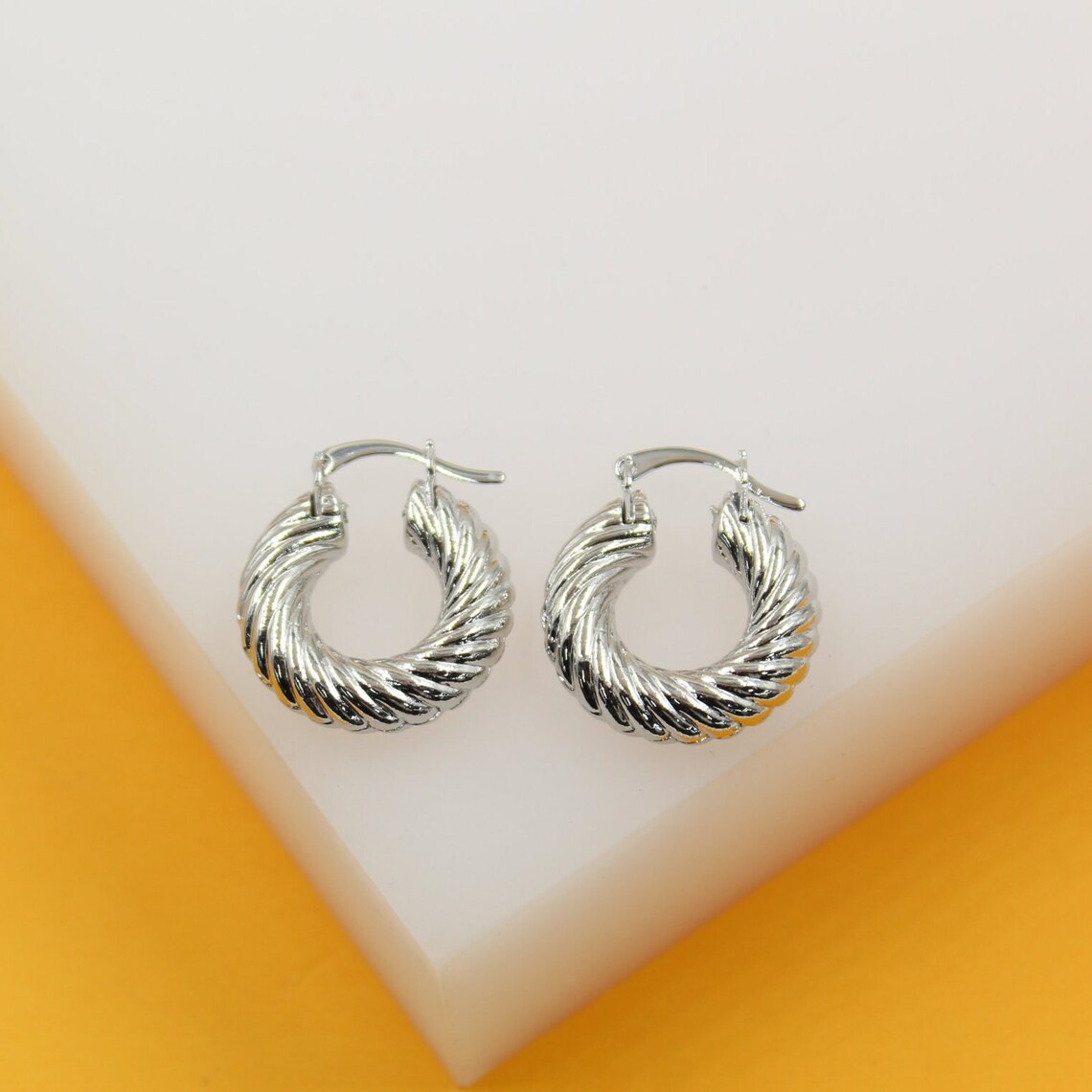 A pair of shiny silver twisted hoop earrings rests on a white and yellow display block.