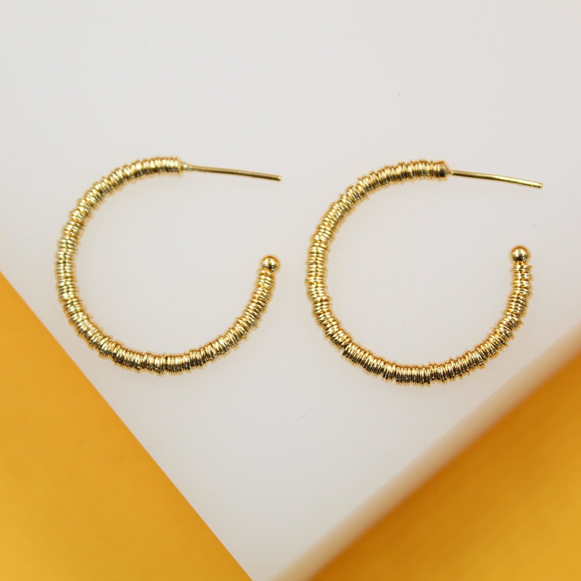 A pair of textured gold hoop earrings rests on a white and yellow surface.