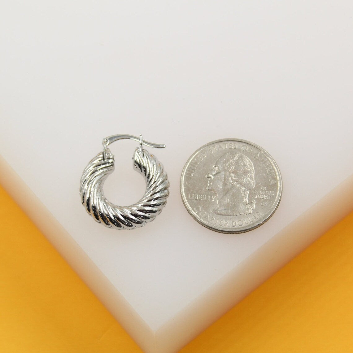 A glossy silver chunky twisted hoop earring rests next to a US quarter on a white display stand for size comparison.