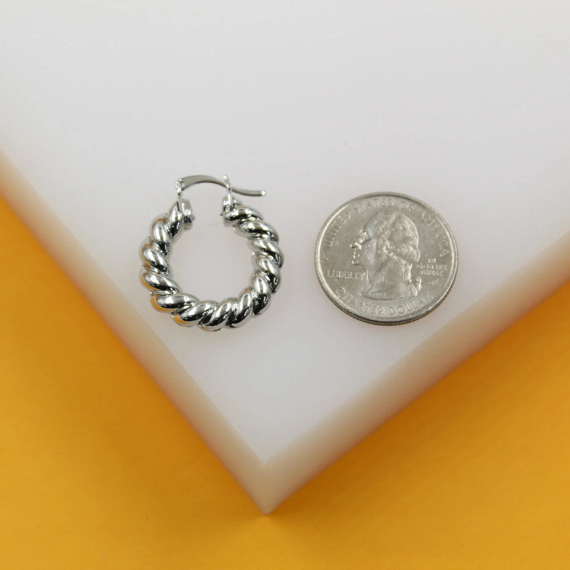 A chunky silver twisted hoop earring sits next to a US quarter coin for scale on a white display surface.