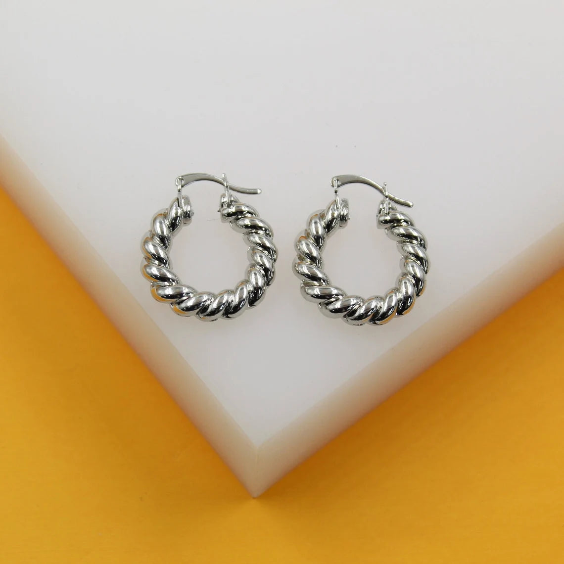 Two polished silver twisted hoop earrings rest side by side on a white display block against a vibrant yellow background.