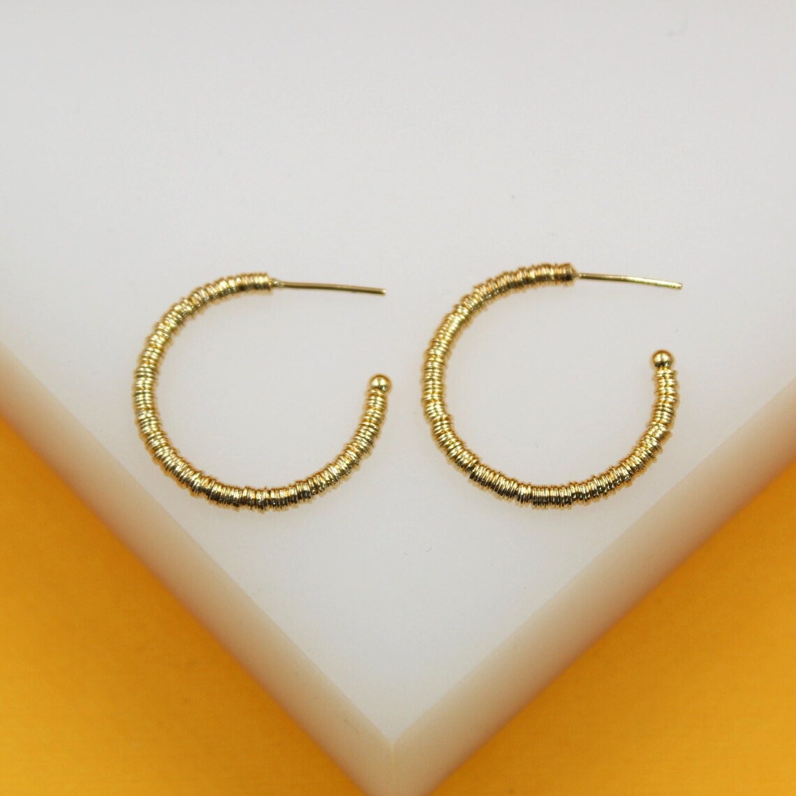 A pair of textured gold hoop earrings rests neatly on a white display surface against a vibrant orange background.