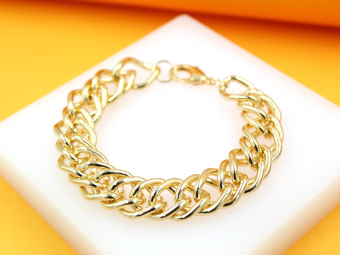 A polished gold chain bracelet rests on a white display block against a vibrant yellow and orange backdrop.
