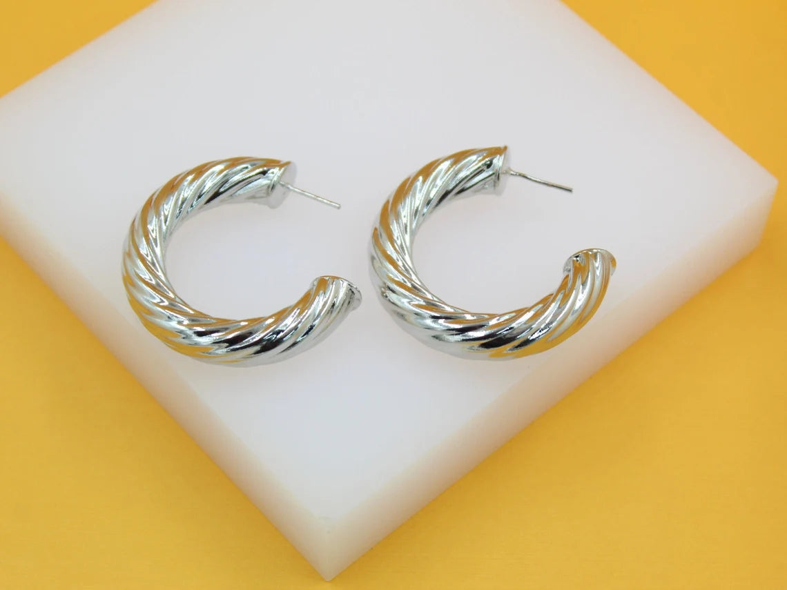 A pair of glossy, silver-plated twisted hoop earrings rests on a white display block against a warm yellow background.