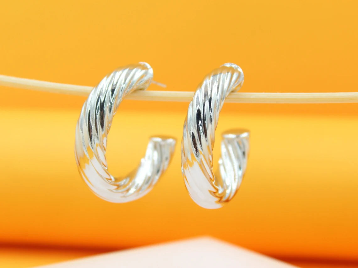 A pair of polished silver twist hoop earrings hangs delicately on a wooden stick against a warm orange background.