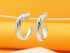 A pair of polished silver twist hoop earrings hangs delicately on a wooden stick against a warm orange background.