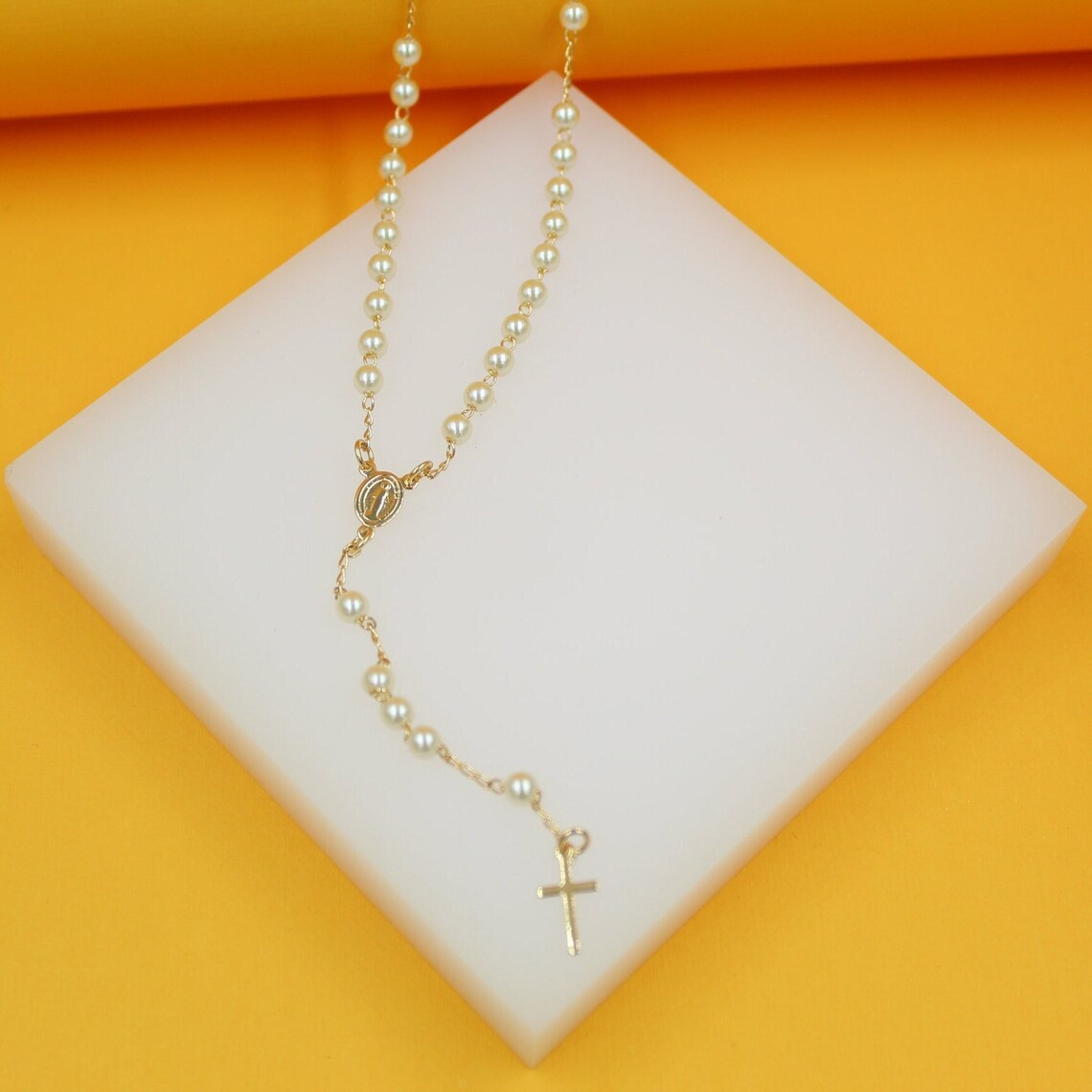 A delicate gold pearl rosary rests gracefully on a white display block against a vibrant yellow background.