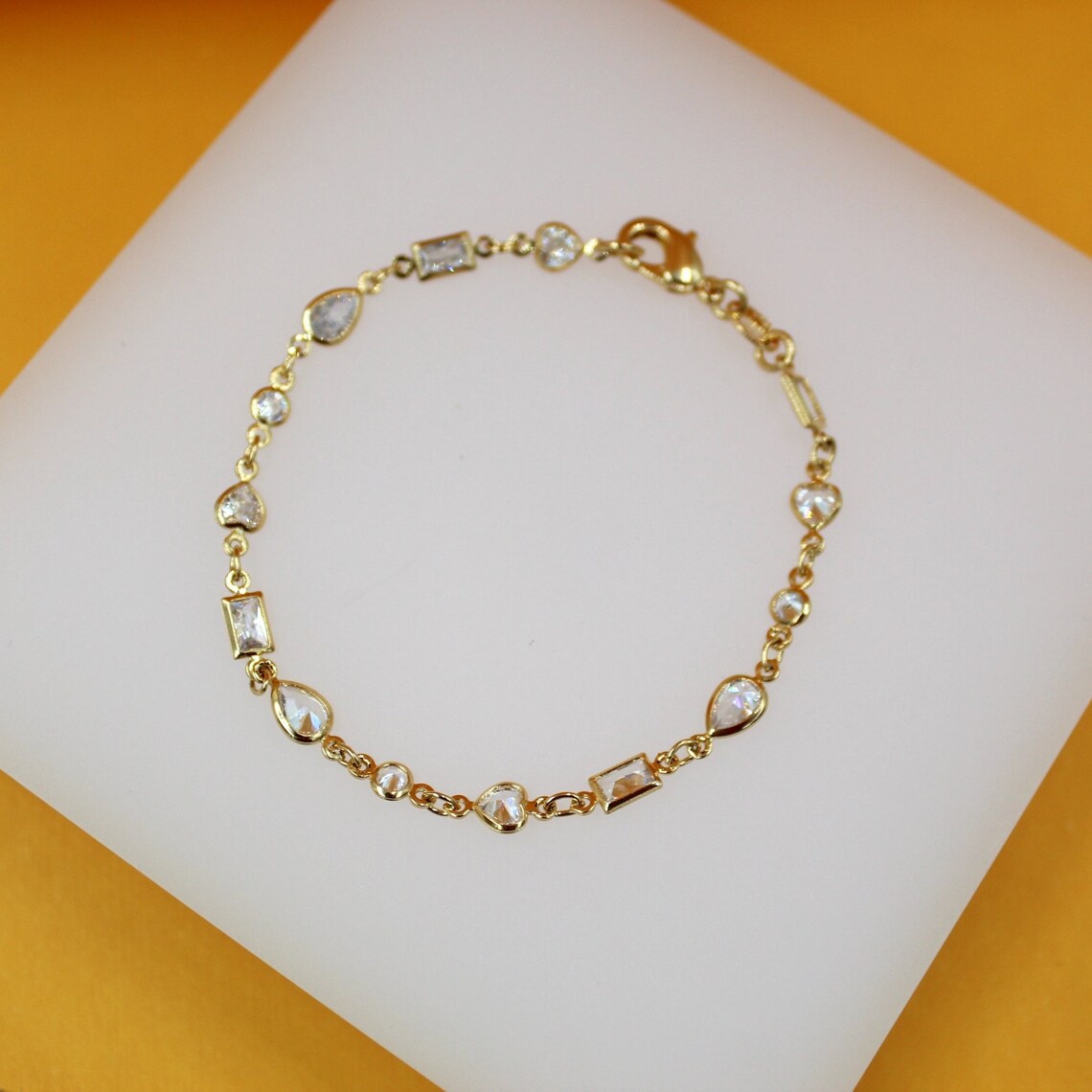A delicate gold charm bracelet featuring mixed geometric gemstone cuts rests on a white display surface against a warm yellow background.