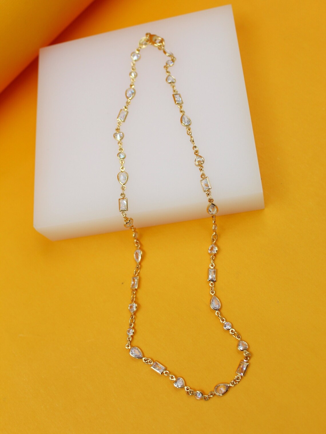 A delicate gold chain necklace featuring alternating round and rectangular crystal stones rests on a white display block against a vibrant yellow background.