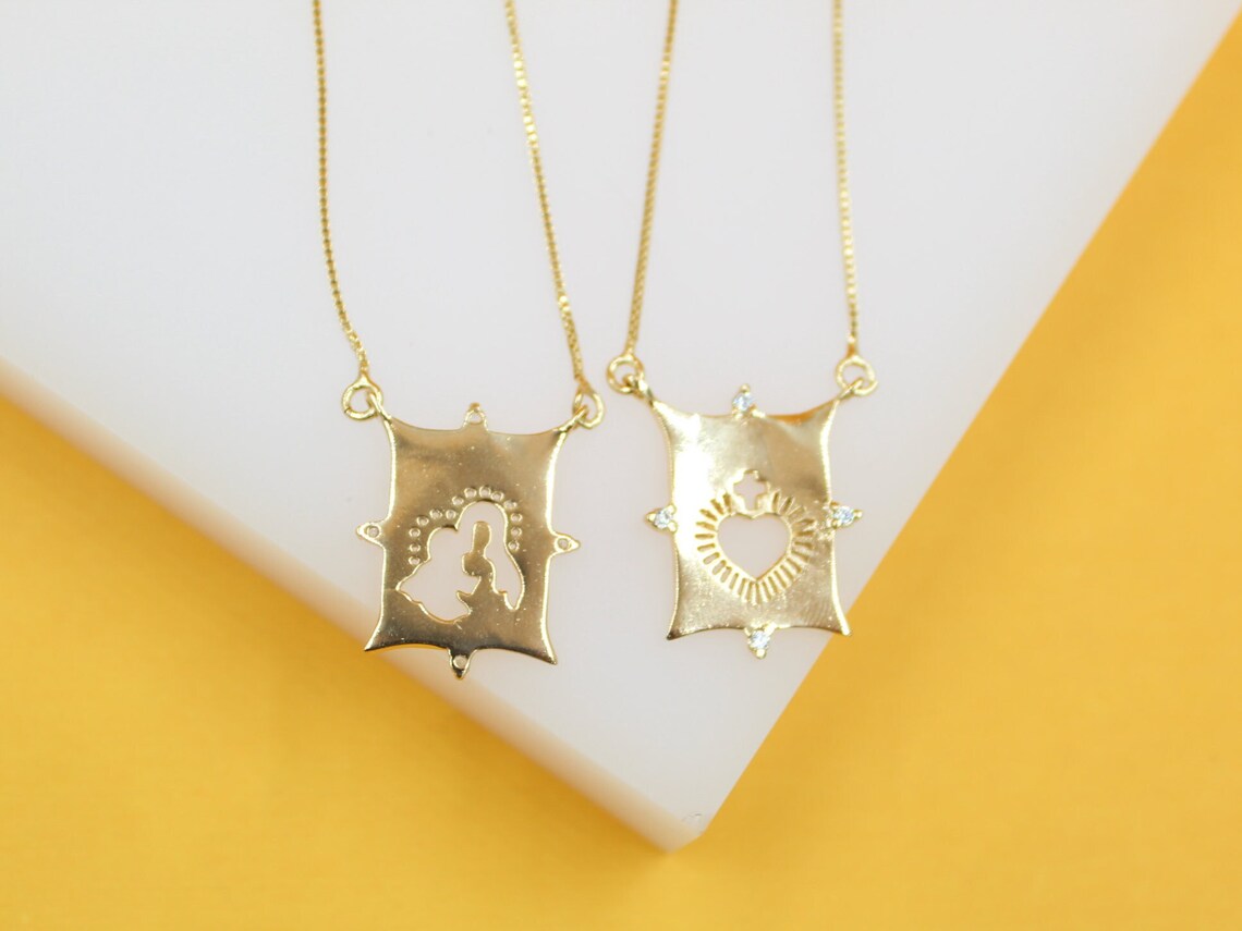 Two gold religious necklaces feature intricate pendant charms resting on a white display against a vibrant yellow background.