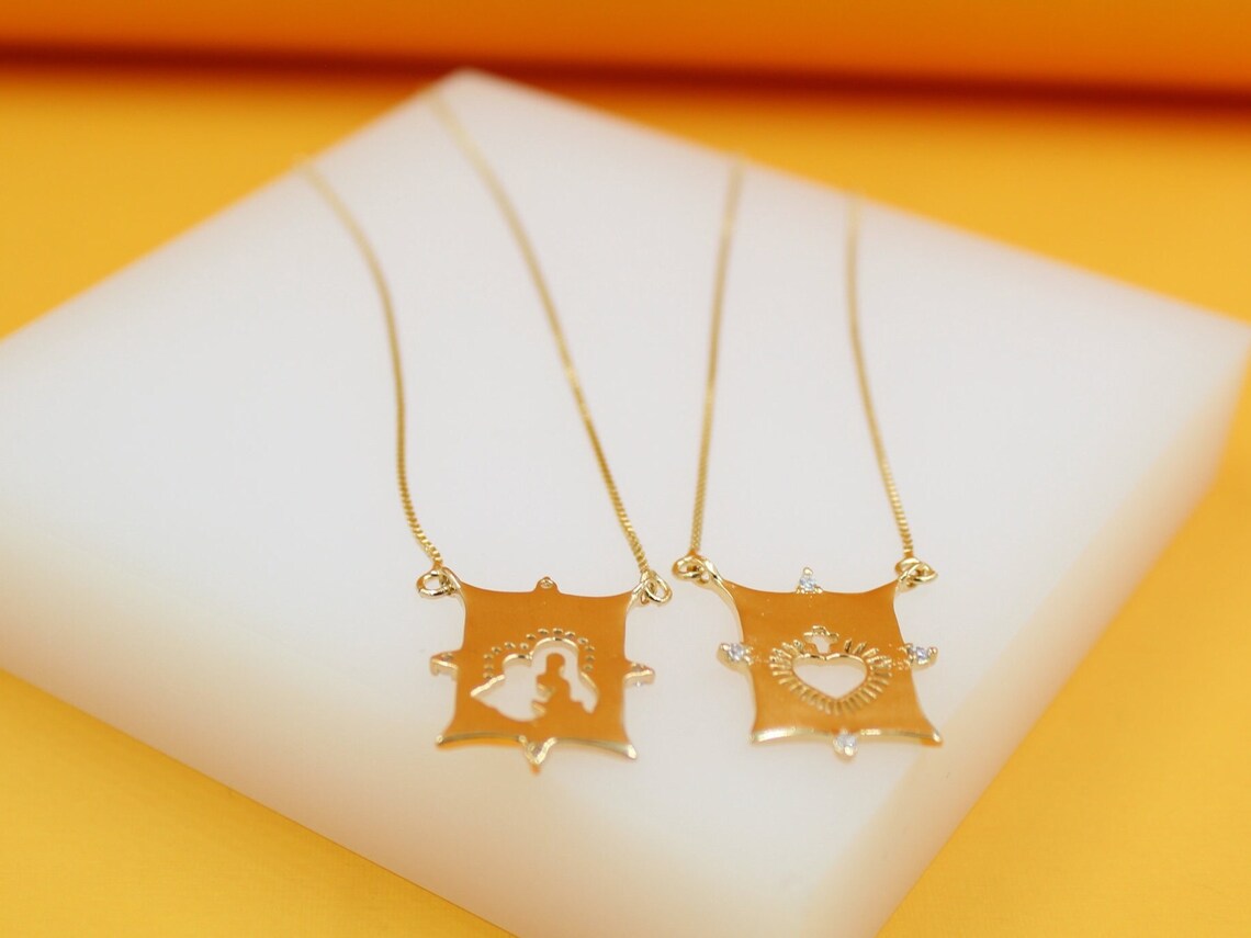 Two gold necklaces featuring square religious pendants rest on a white display block against a warm yellow background.