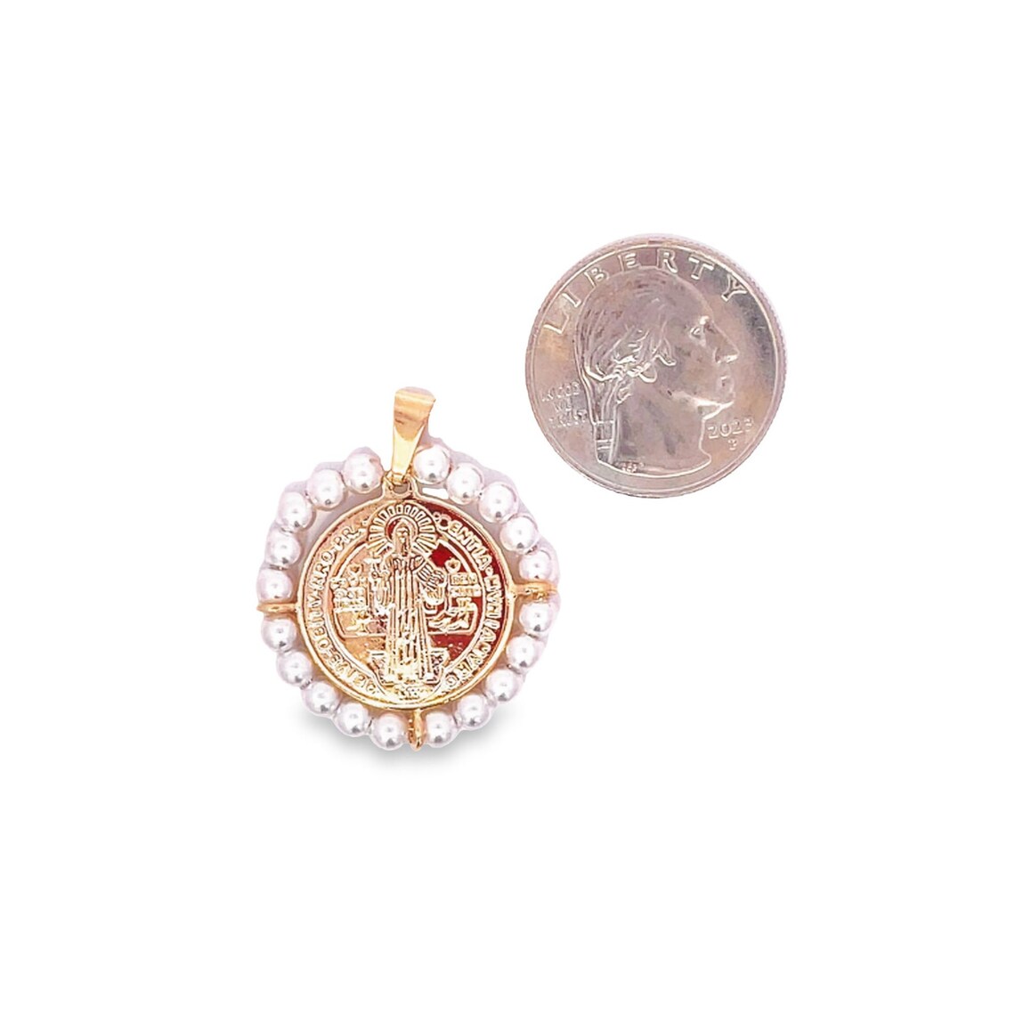 A gold Saint Benedict medallion pendant encircled by small white pearls rests next to a US quarter coin for scale.