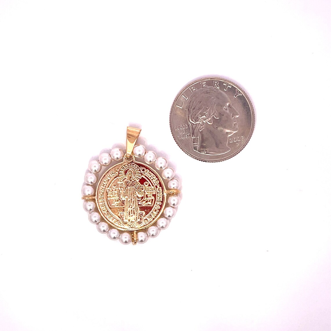 A gold Saint Benedict medal pendant framed by a halo of small white pearls rests next to a US quarter for scale.