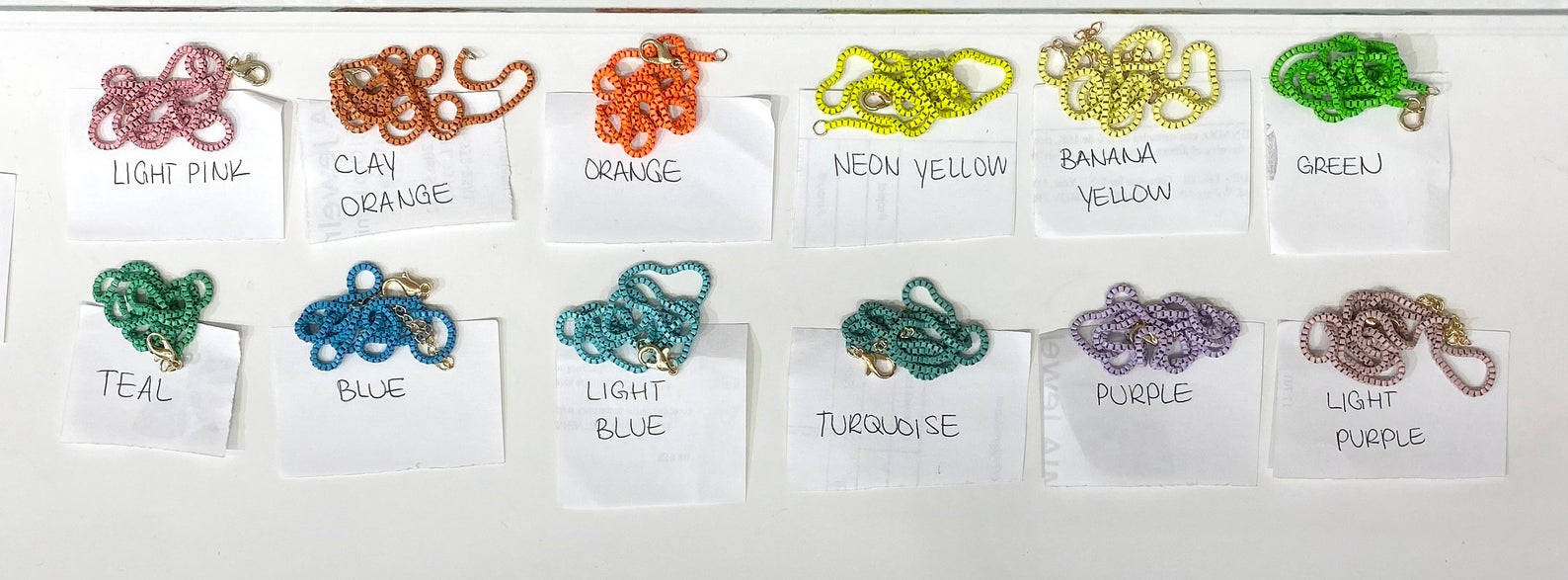 A top-down view displays twelve colorful, tiled box-chain necklaces arranged neatly on white paper labels with handwritten color names.