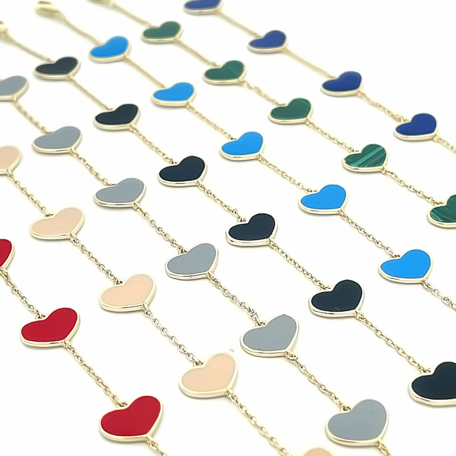 Multiple gold chain bracelets feature colorful enamel heart charms in shades of red, beige, grey, black, blue, and green, arranged neatly on a white surface.