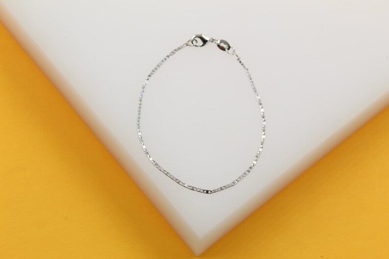 A delicate silver chain bracelet rests neatly on a white and yellow display block.