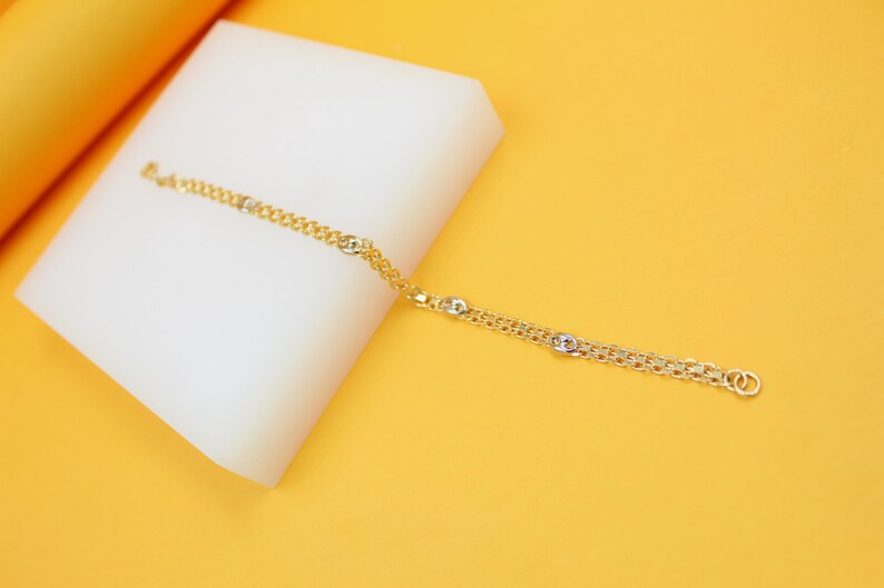 A delicate gold chain bracelet with interspersed silver links rests elegantly on a white display block against a vibrant yellow background.