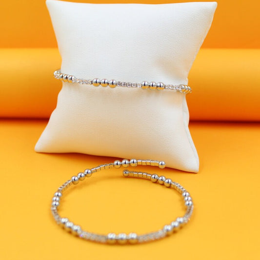 A shiny silver beaded bracelet rests on a white display pillow against a vibrant orange background, with another matching spiral bracelet displayed below it.