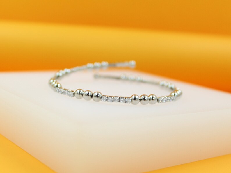 A delicate silver charm bracelet featuring alternating polished metal beads and sparkling cubic zirconia stones rests elegantly on a white display stand against a warm yellow background.