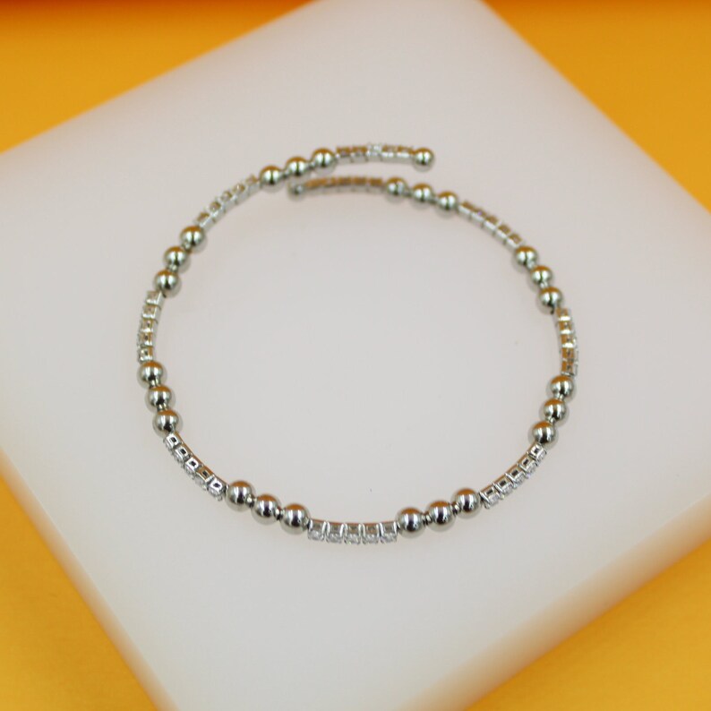 A silver spiral bangle bracelet featuring alternating polished metal beads and cubic zirconia stones rests on a white display surface.