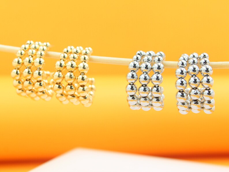 Two pairs of gold and silver beaded hoop earrings rest on a wooden stick against a vibrant yellow backdrop.