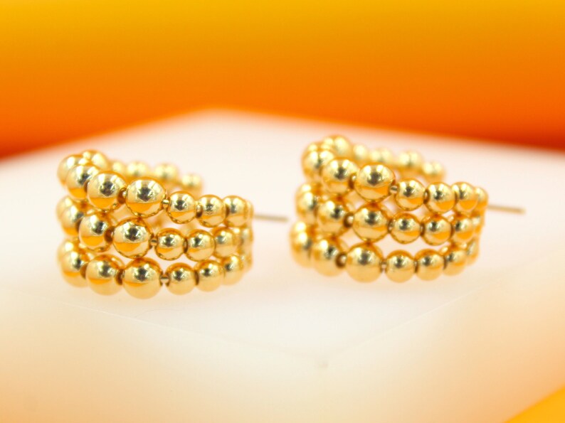 A pair of multi-row beaded gold hoop earrings rests on a reflective white surface against a warm orange backdrop.